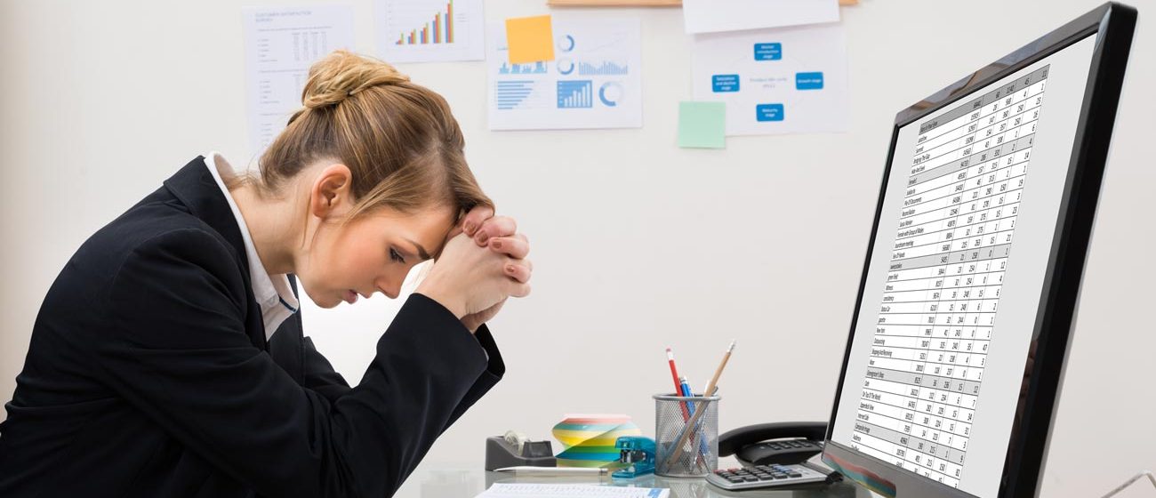 Stressed businesswoman struggling with Excel Spreadsheets for her HR analytics shown at her desk in front of a computer with charts hanging in the background