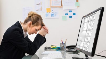 Stressed businesswoman struggling with Excel Spreadsheets for her HR analytics shown at her desk in front of a computer with charts hanging in the background