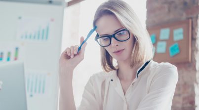 HR professional thinking about her retention strategy while reviewing HR data and analytics with glasses on and a pen in her office